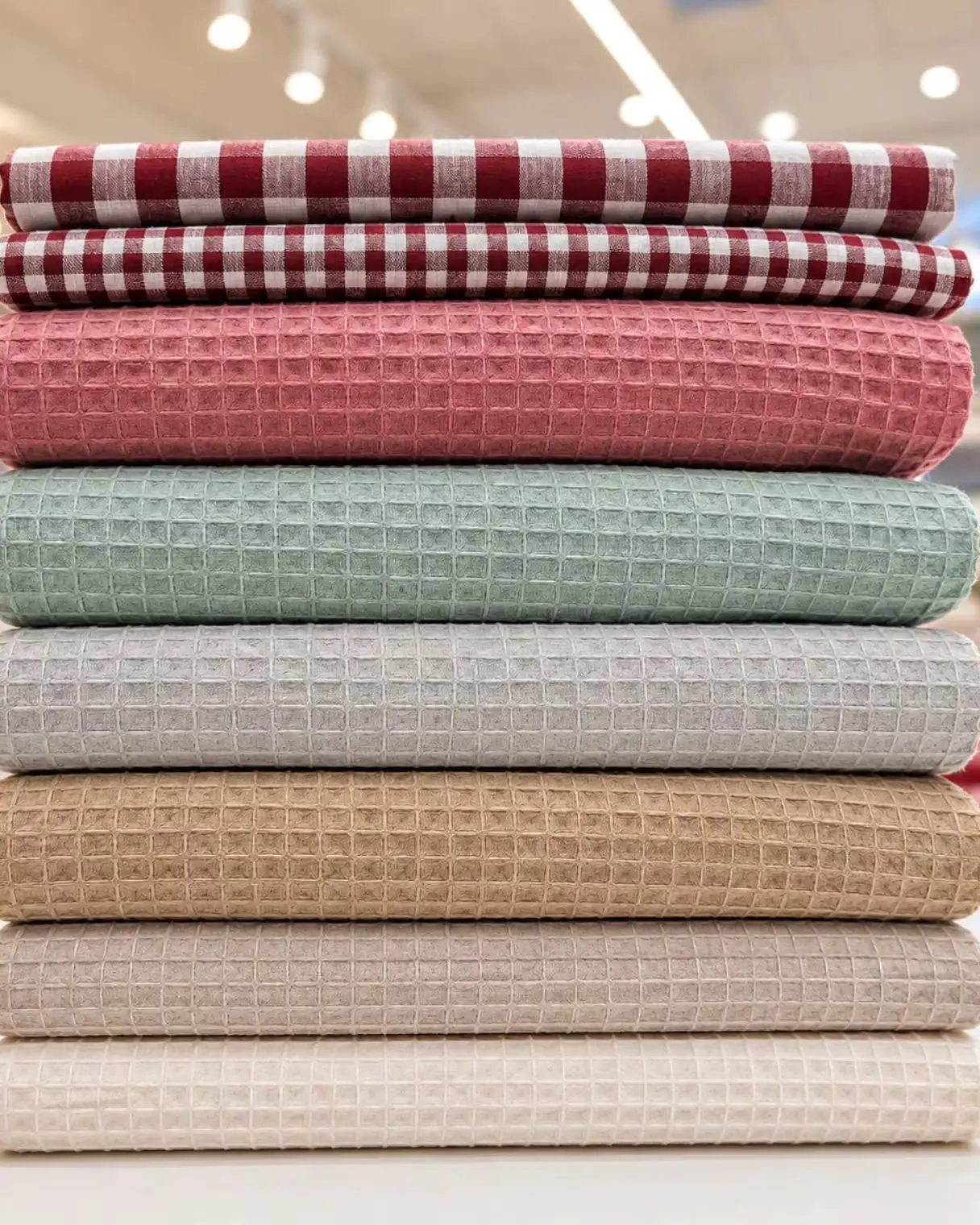 Pila de telas waffle de algodón en rosa, verde agua, gris, beige y crema, con dos cuadros vichy arriba, en el local de Clementelas